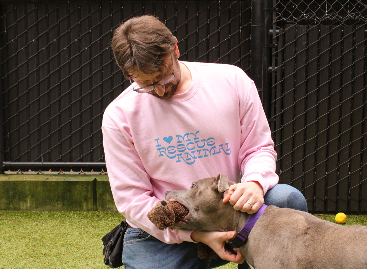 An image of a man wearing an I heart my rescue animal sweatshirt playing with a dog