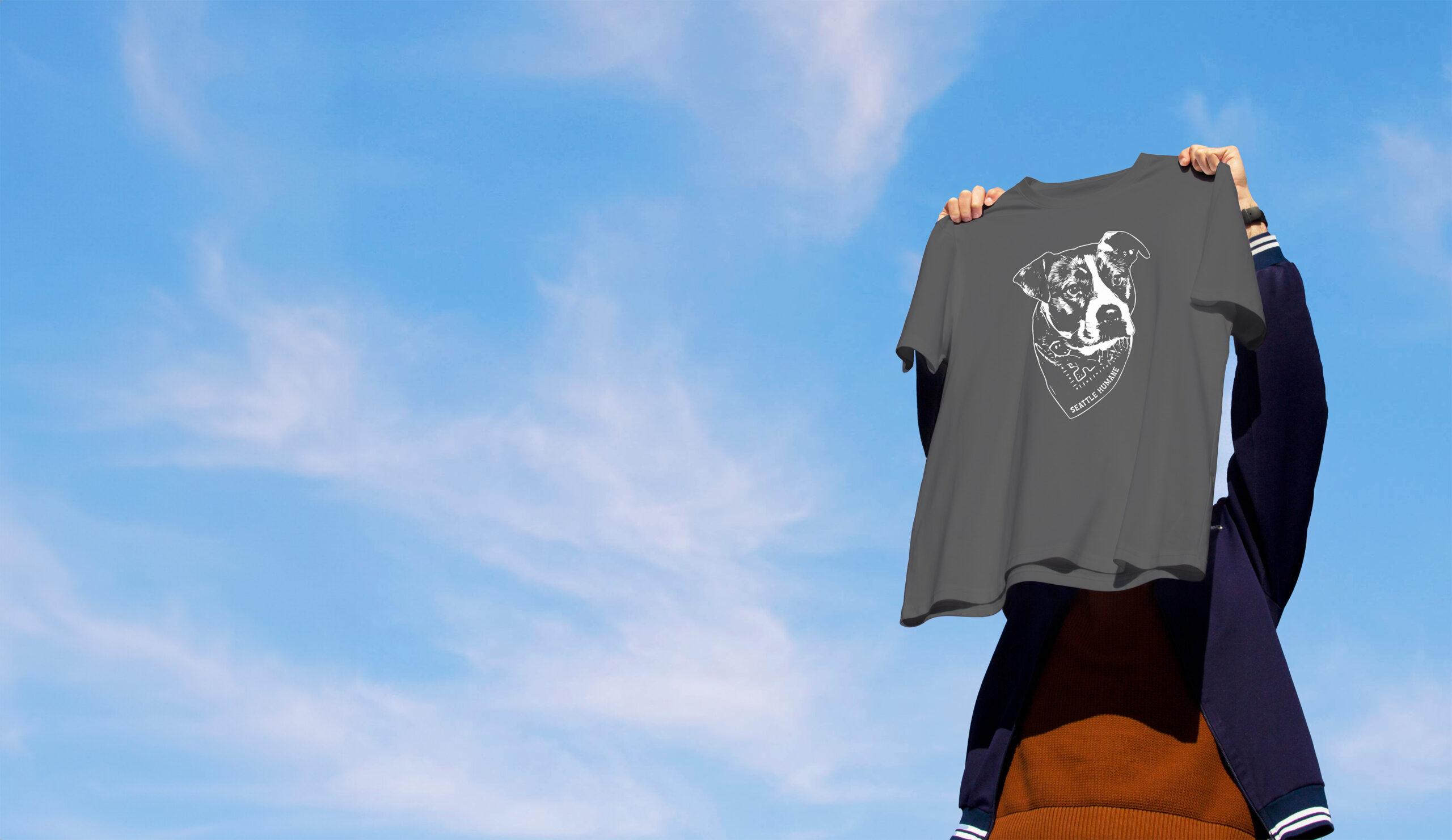 A person on a blue sky background holds up a grey t-shirt with a dog on it.